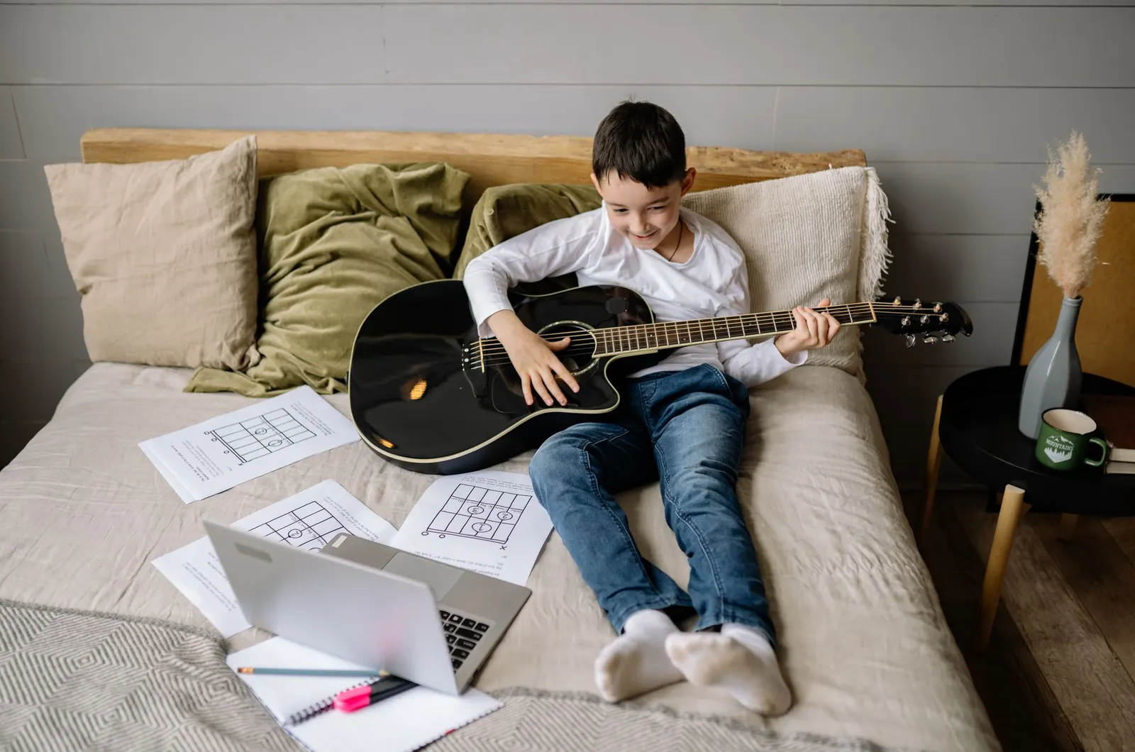 Boy playing acoustic guitar on his bed with a laptop and music sheets